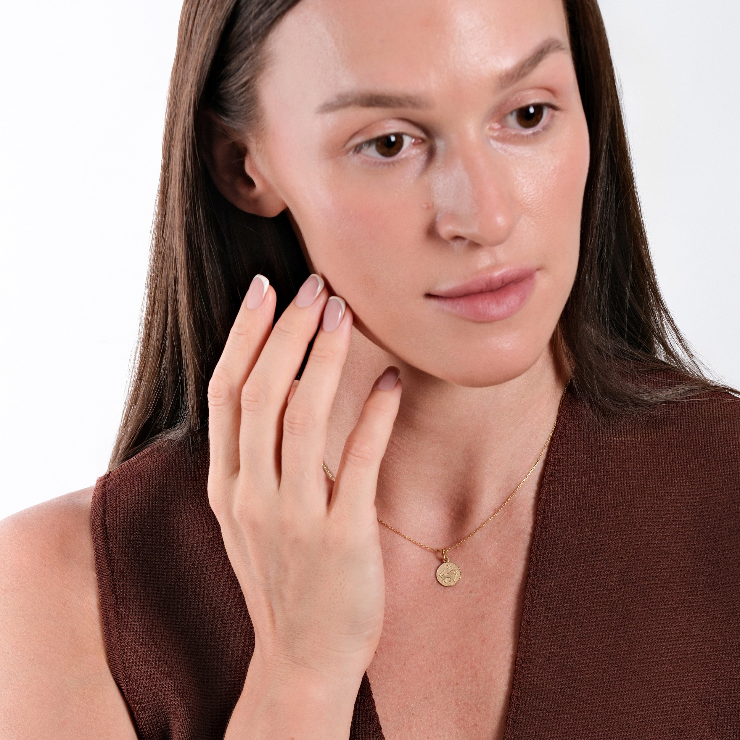 Woman with natural makeup wearing gold necklace and brown top, touching face gently with manicured hand.