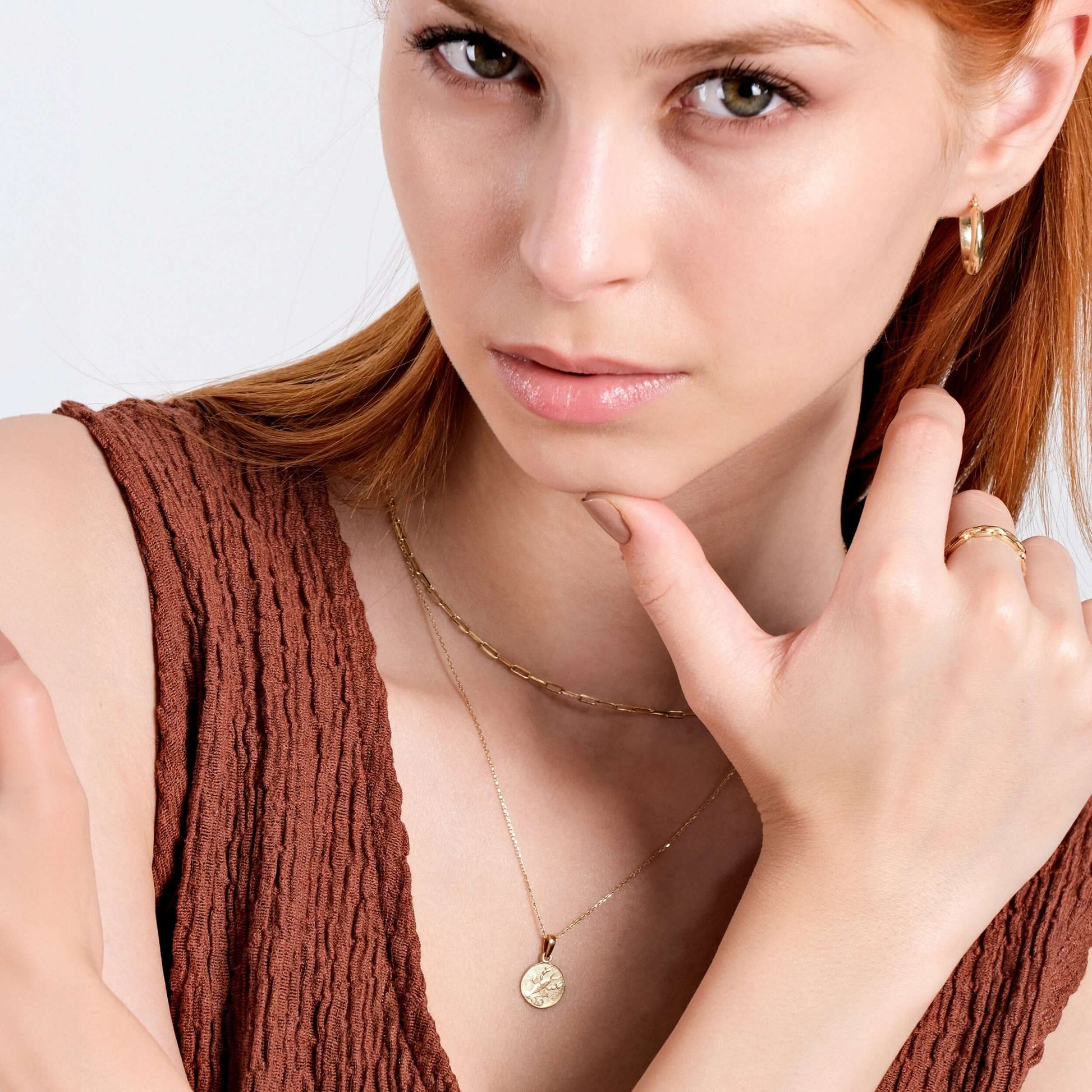 A woman with red hair, dressed in a sleeveless textured brown top, poses with her hand touching her face. She is adorned with gold hoop earrings and a delicate 14K solid gold ring. Around her neck, she wears layered necklaces featuring the Mionza Scorpio Zodiac Necklace made of 14K solid gold. The neutral background accentuates her minimalist style.