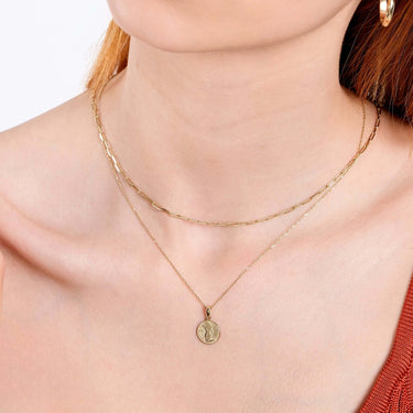 Close-up of a person wearing two gold necklaces. The top necklace features a simple chain, while the bottom one is the Mionza Virgo Zodiac Necklace in 14K Solid Gold with an engraved design. The person is wearing a sleeveless red top and has light-colored hair—an elegant birthday gift idea for Virgos.