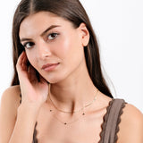 Woman with long hair wearing layered necklaces and a brown top, touching her face, against white background.