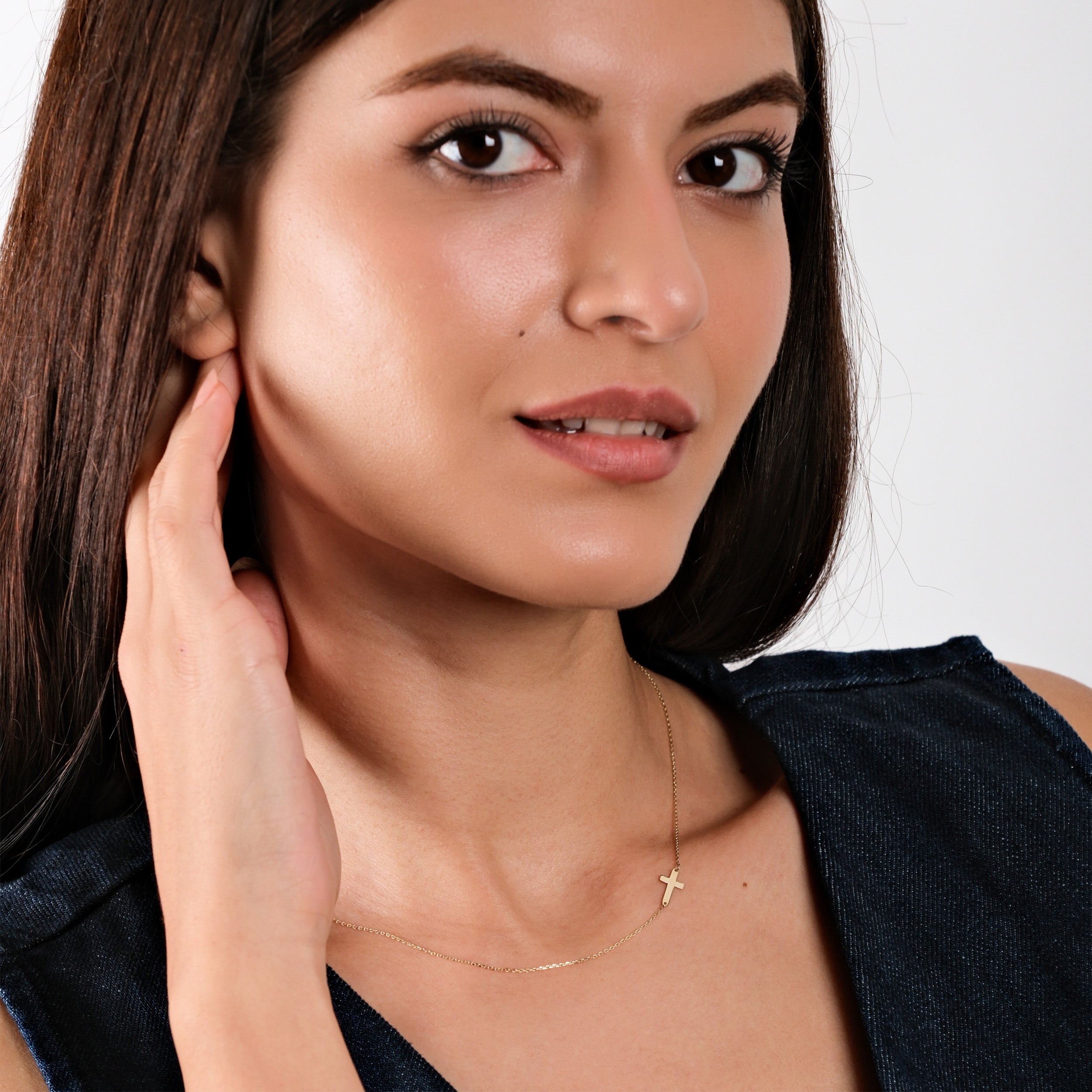 Woman with long brown hair wearing a gold cross necklace and denim top, posing elegantly.