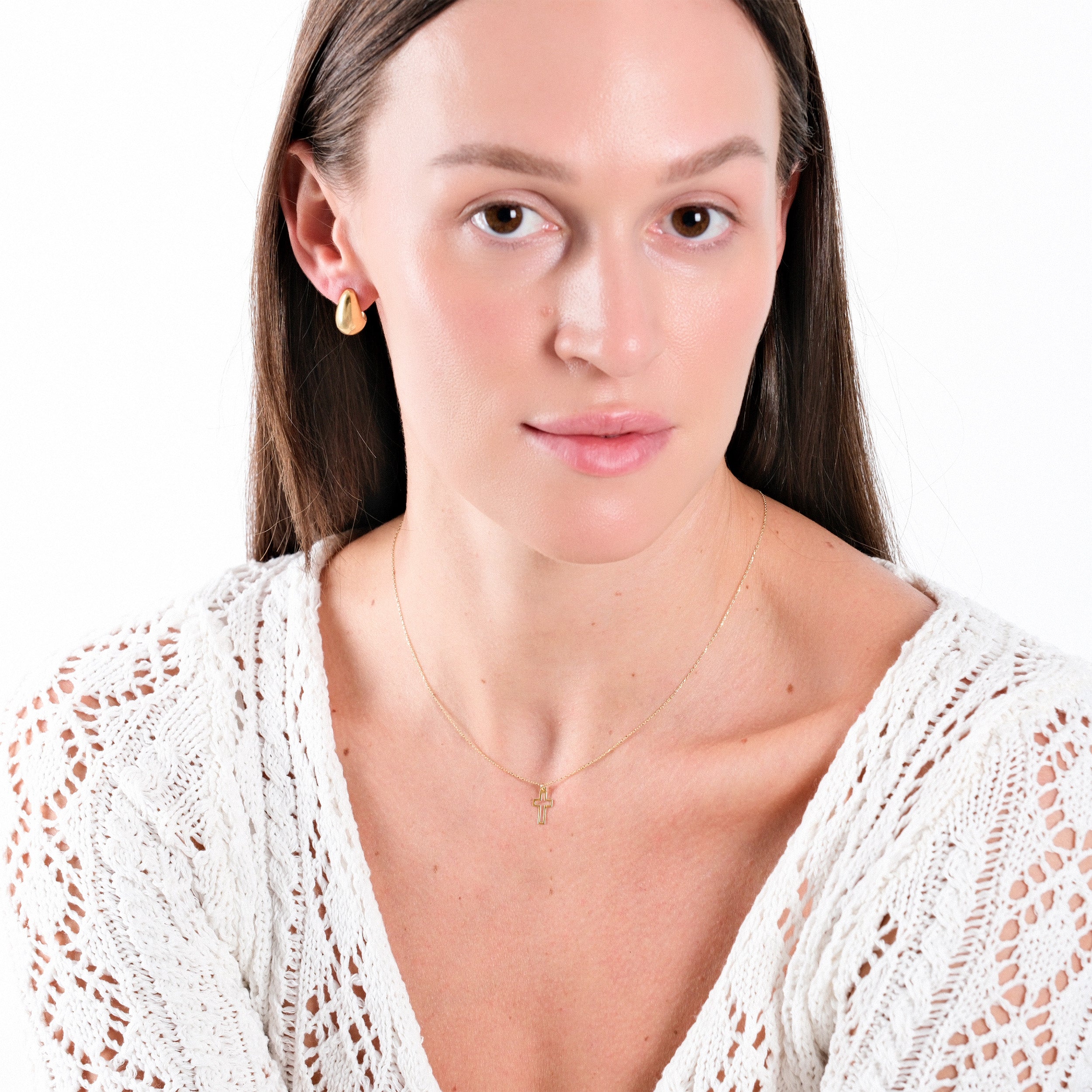 Woman wearing gold earrings and necklace, dressed in white crochet top, on a plain background. Minimalist fashion.