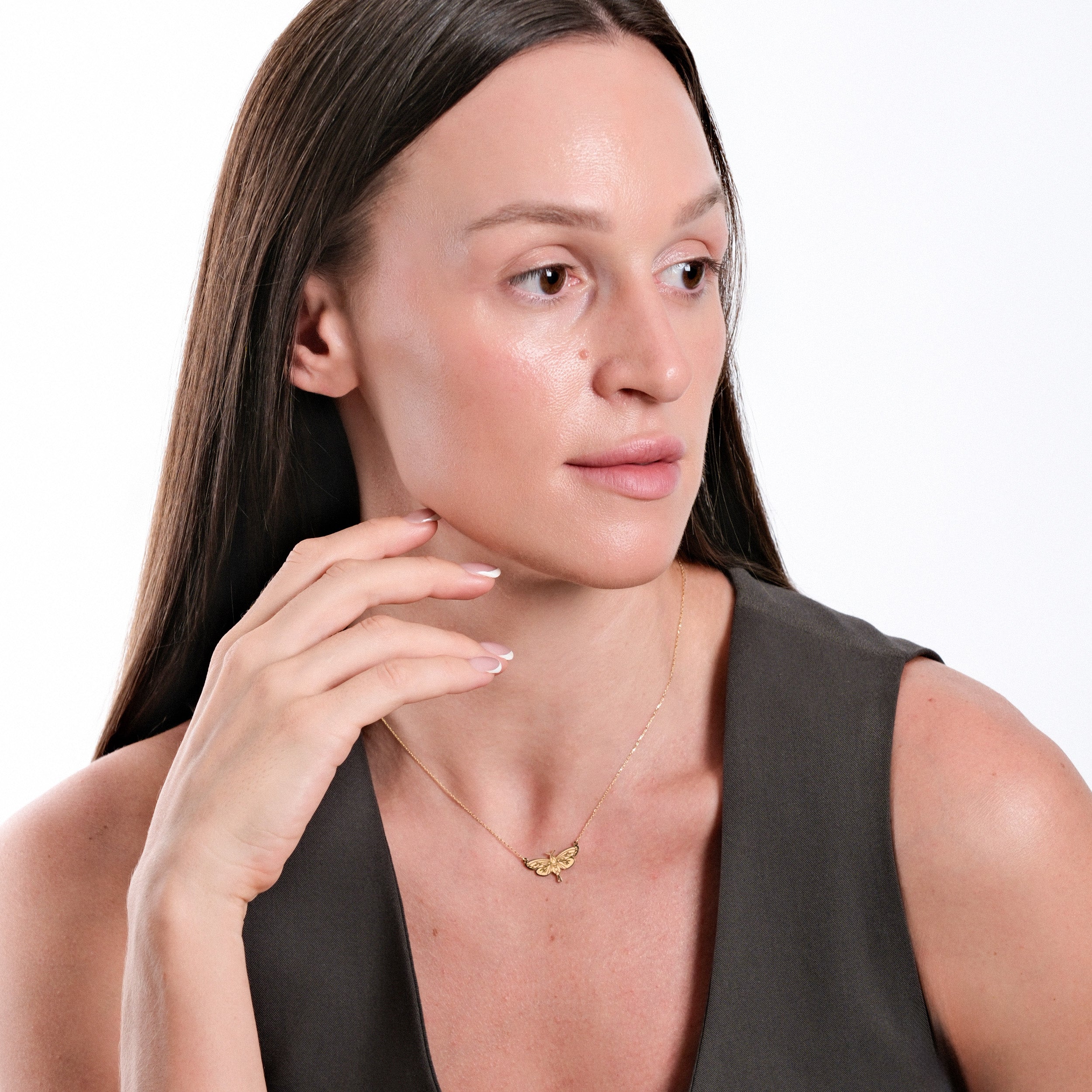 Woman with long hair wearing a butterfly necklace, gazing sideways, in a sleeveless top against white background.