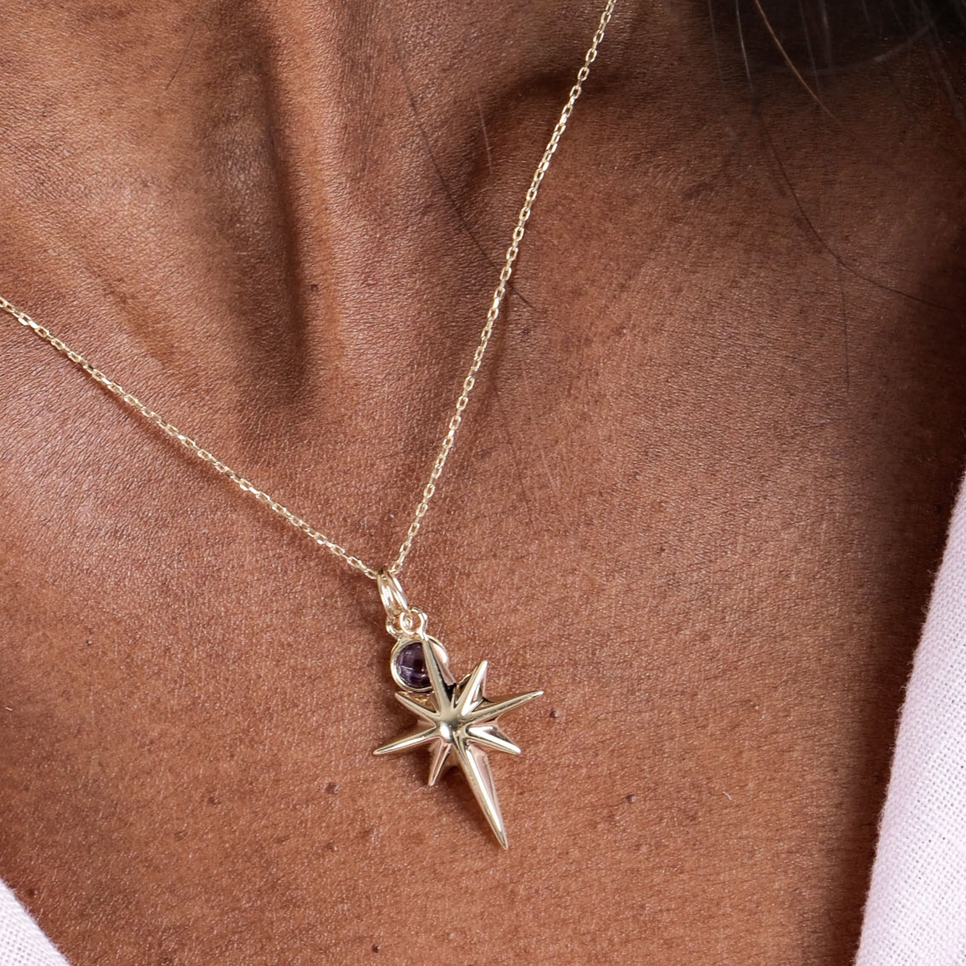 Gold star pendant necklace with amethyst accent on a delicate chain, worn on a brown skin neckline.