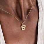 Gold necklace with letter E and small charm pendant on person's neck, wearing a striped shirt.