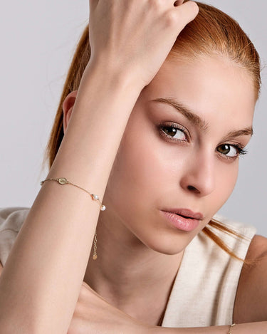 A woman with fair skin and auburn hair gazes at the camera, her head resting on her bent arm. She wears a sleeveless beige top and a delicate Mionza Miraculous Medal Bracelet with Real Pearl in 14K Solid Gold adorned with a small charm and pearl. The background is a simple light gray.