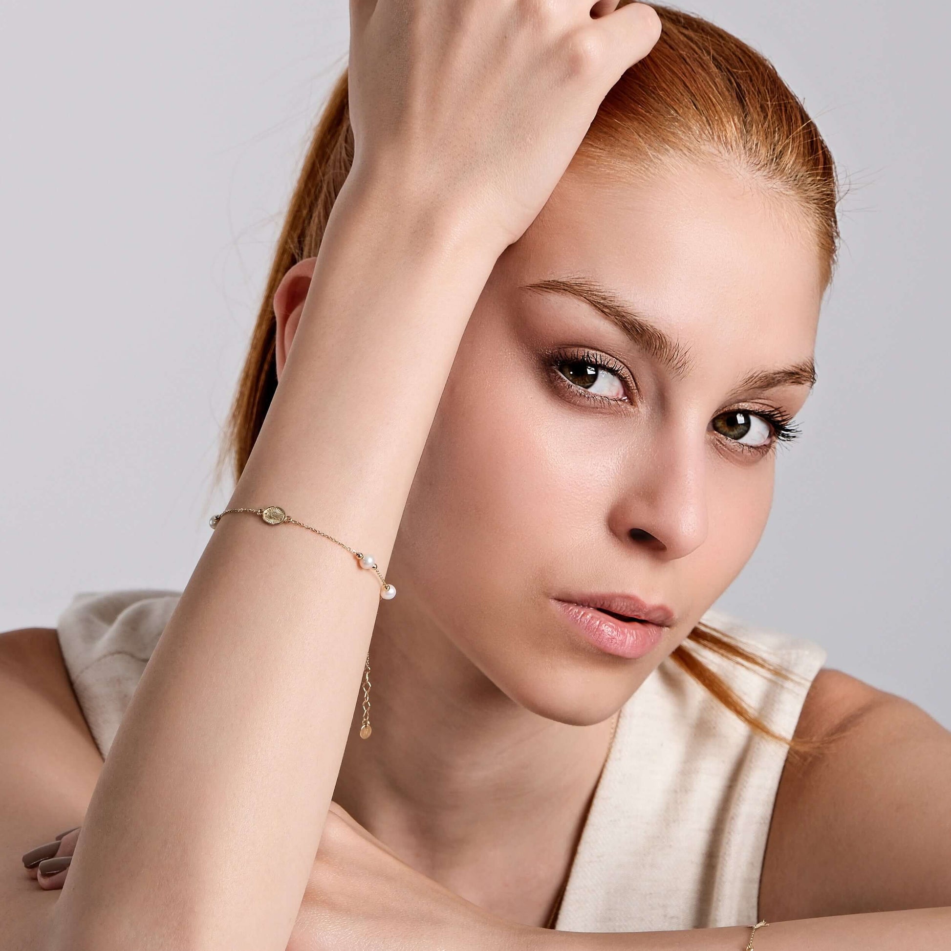 A woman with fair skin and auburn hair gazes at the camera, her head resting on her bent arm. She wears a sleeveless beige top and a delicate Mionza Miraculous Medal Bracelet with Real Pearl in 14K Solid Gold adorned with a small charm and pearl. The background is a simple light gray.