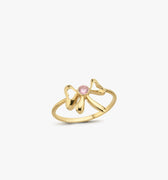 The Mionza Bow Ring in 14K Solid Gold showcases a delicate bow design adorned with a pink gemstone set at its center. The band's textured pattern enhances the overall elegance of this knot love ring, all highlighted against a plain white background to emphasize the intricate details.