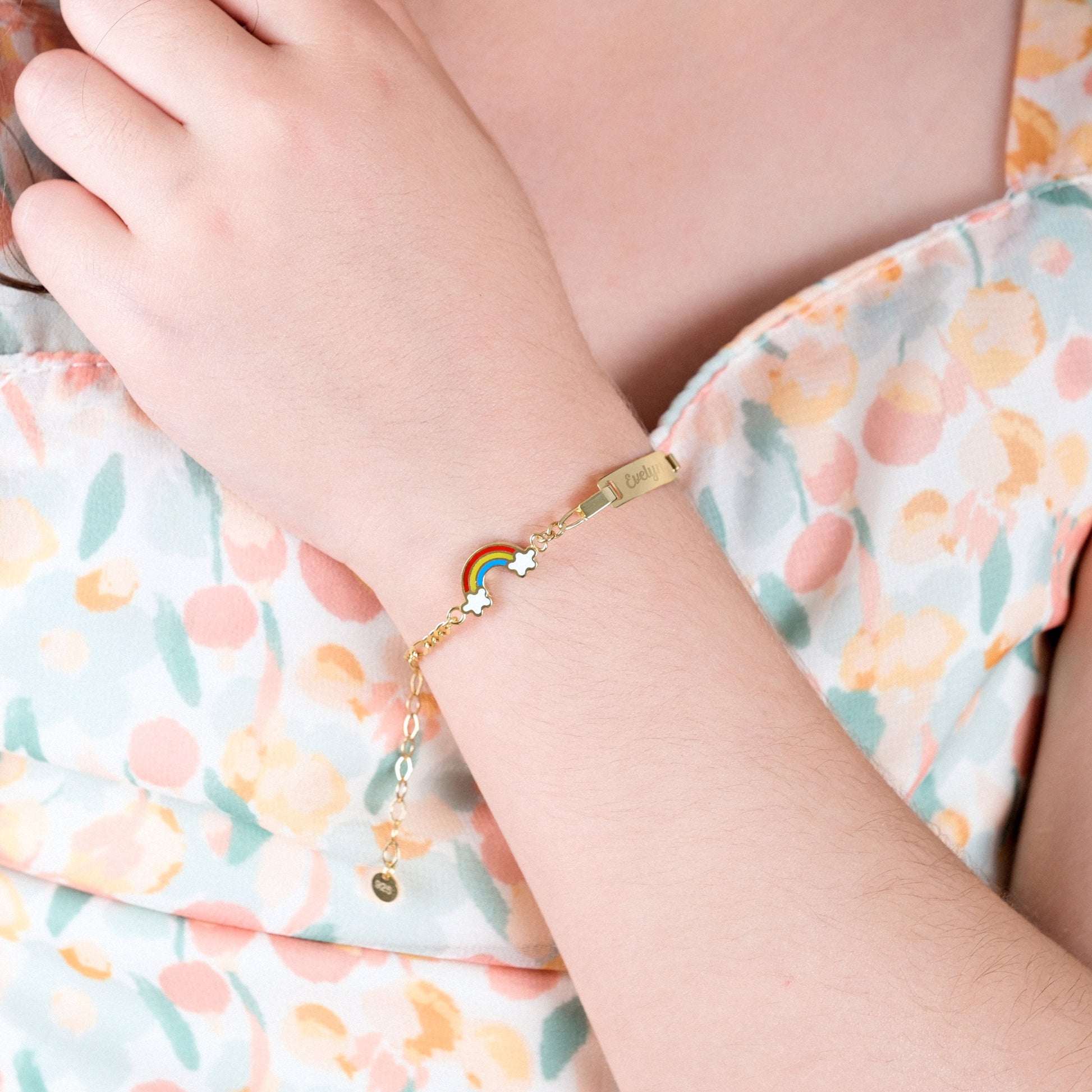 Child's arm wearing a gold baby bracelet with rainbow and star charms, against a floral background.