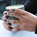 Tattooed hands holding a matcha latte in a ceramic mug, adorned with dark nails and a gold ring.