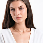 Woman wearing layered gold necklaces with initial pendant, white blouse, posing against a plain background.