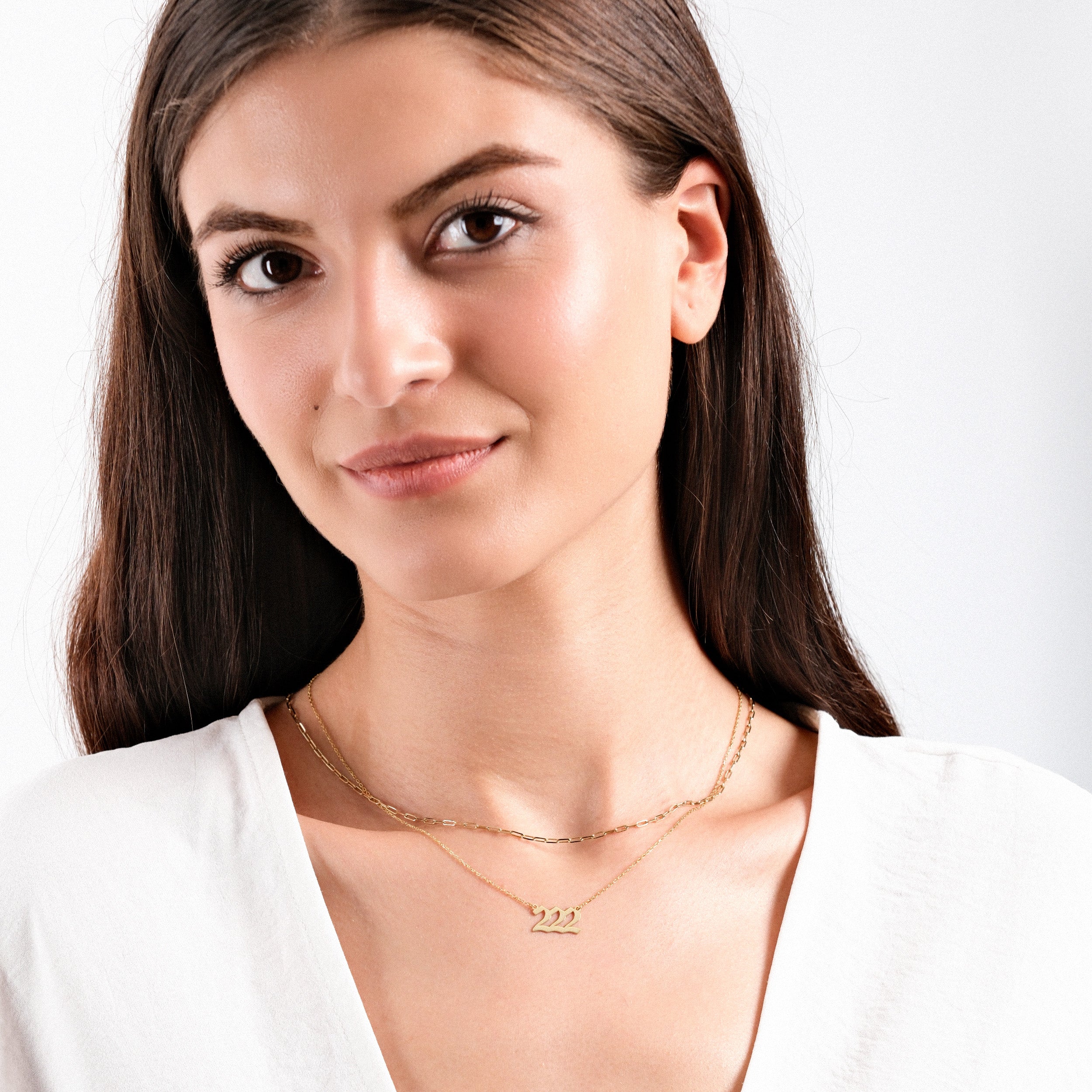 Woman wearing delicate gold necklace with number 222 pendant, white blouse, smiling gently, neutral background.