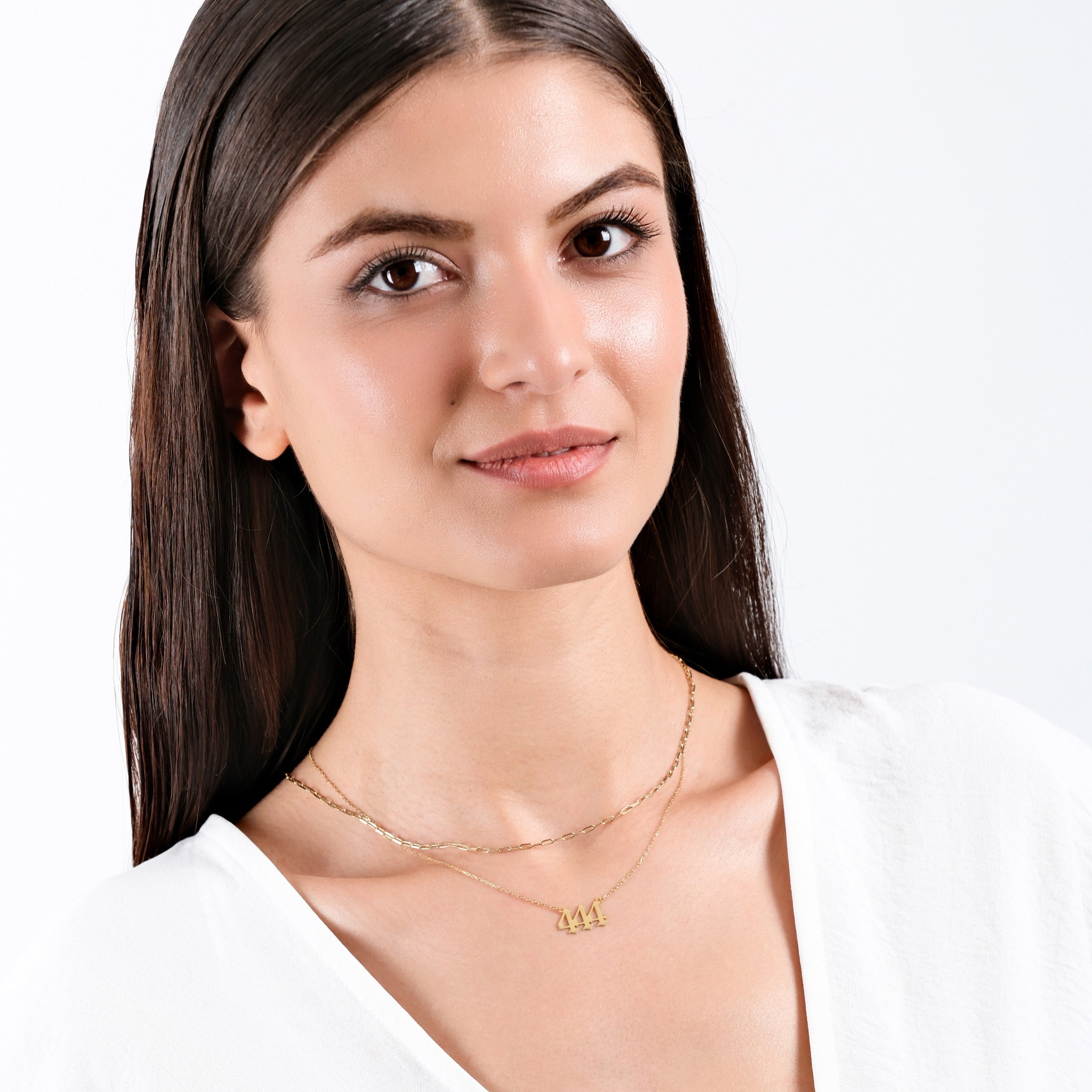 Woman in white shirt wearing layered gold necklace with "444" pendant, smiling confidently against a white background.