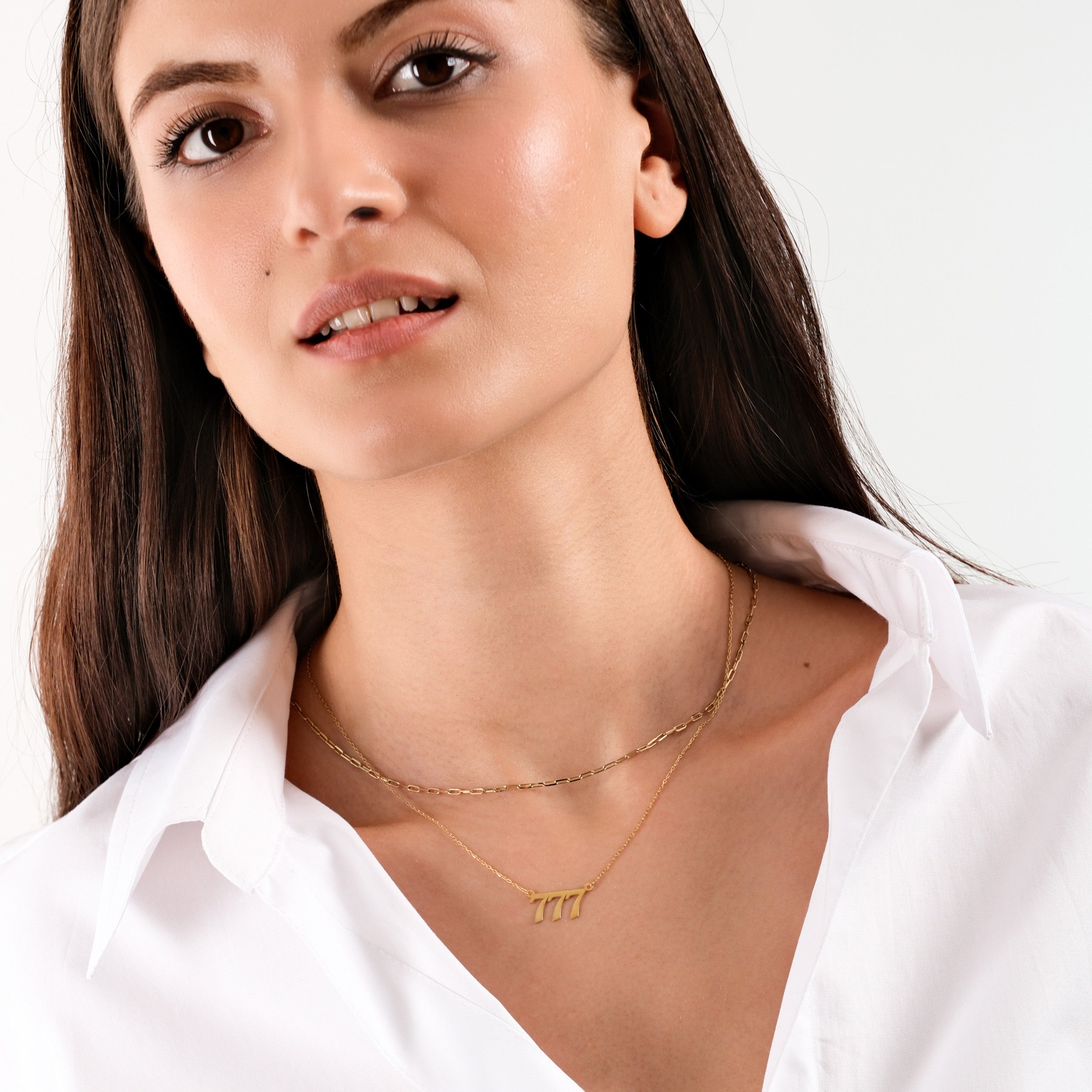 Woman wearing a white shirt and layered gold necklaces, including a "777" pendant, against a neutral background.