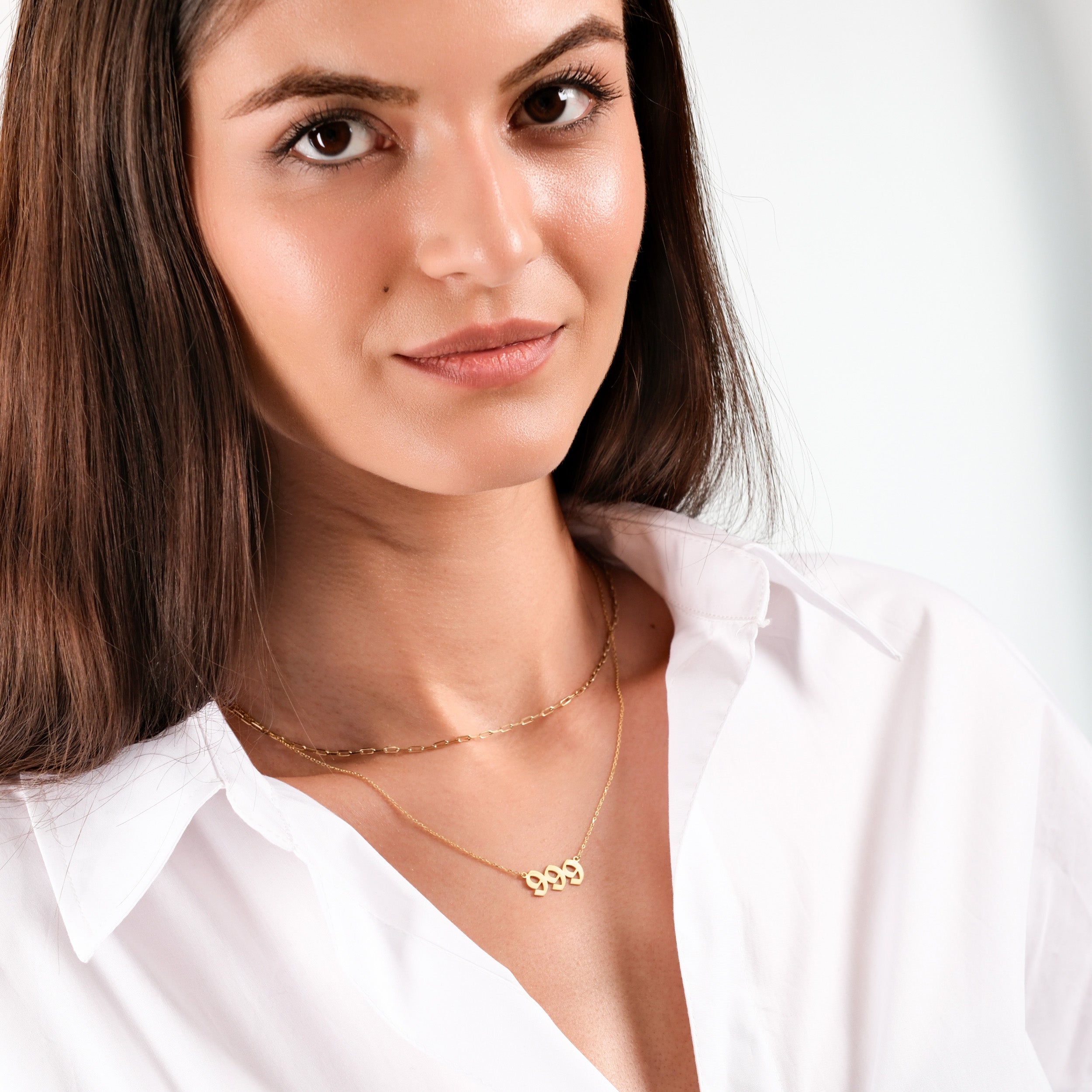 Woman in white shirt wearing elegant gold necklaces, smiling confidently against a neutral background.