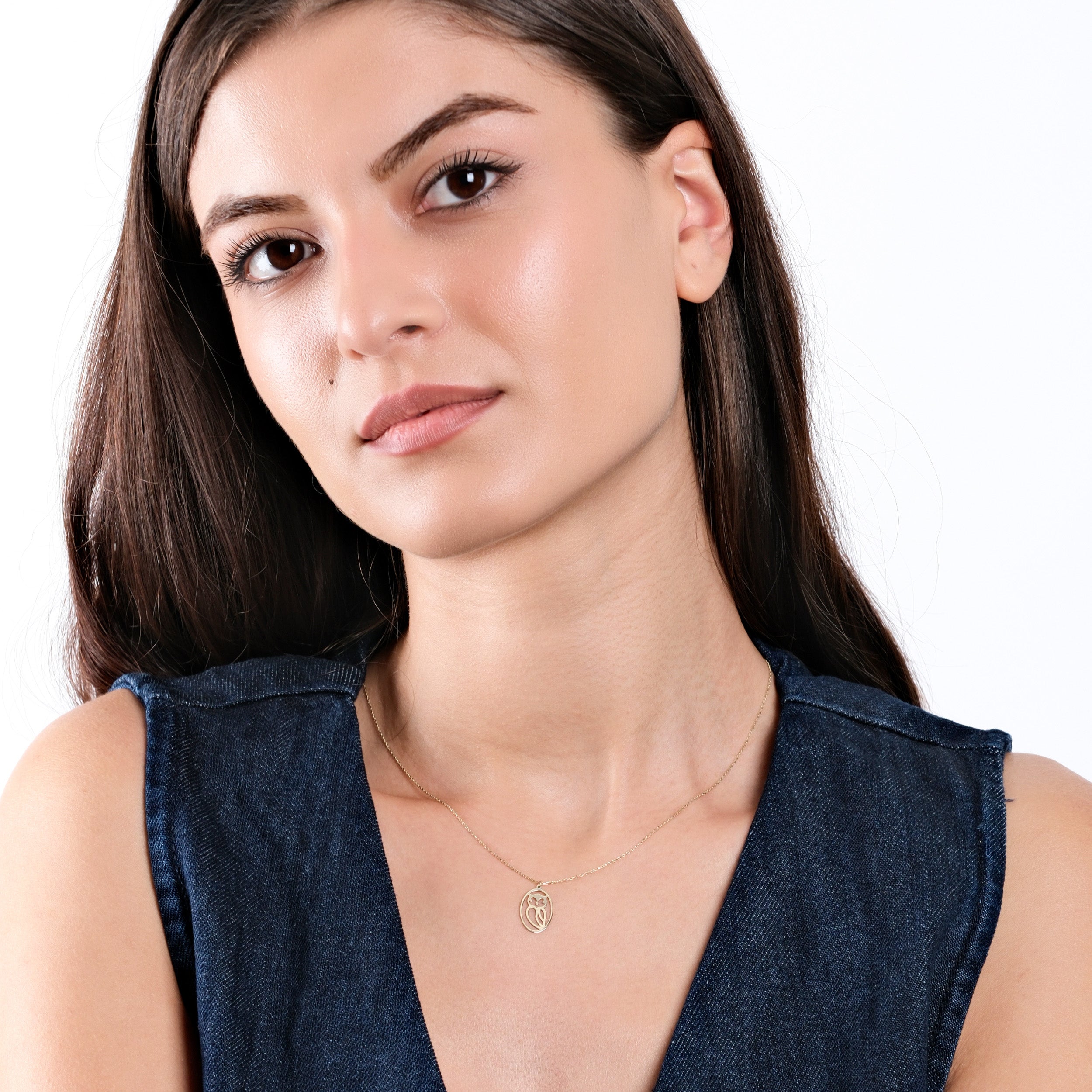 Woman wearing sleeveless denim top and gold necklace, posing against a white background with long, dark hair.