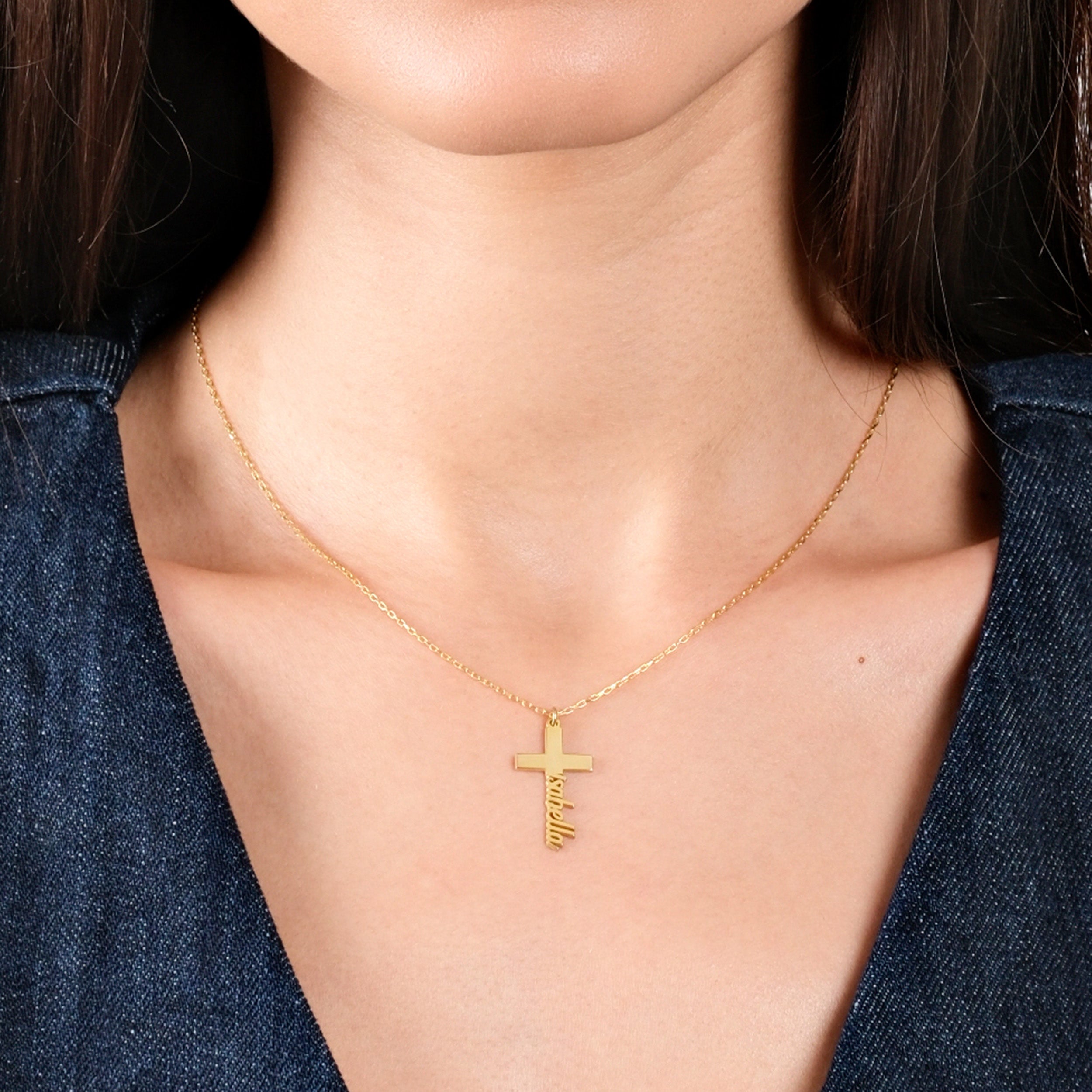 Gold cross necklace with personalized name pendant on woman's neck, wearing a denim top, close-up view.