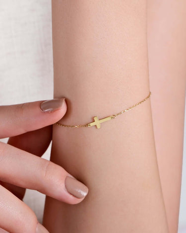 A close-up of a person wearing the Gold Cross Bracelet by Mionza Jewelry, crafted from 14K solid gold with a small sideways cross charm. The person's fingers gently touch the bracelet, showcasing their nude-colored nail polish against a beige, textured top, exemplifying modern faith jewelry at its finest.