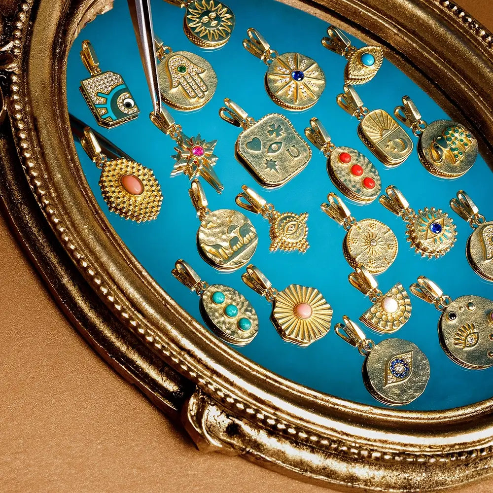 A collection of gold pendants is displayed on a blue background within an ornate, gold-framed setting. The pendants feature various designs, including eyes, hands, hearts, stars, and sunbursts. Some are adorned with gemstones such as turquoise, coral, and pearls. A pair of tweezers is used to hold one of the pendants. The arrangement highlights the intricate details and diversity of the pendants.