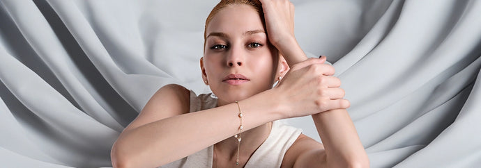 A young woman with light-colored hair poses with her arms crossed in front of her, one hand resting on her head. She wears a sleeveless light-colored top and a gold rosary bracelet with small pearls on her left wrist. Her expression is calm and confident. The background consists of softly draped light gray fabric.