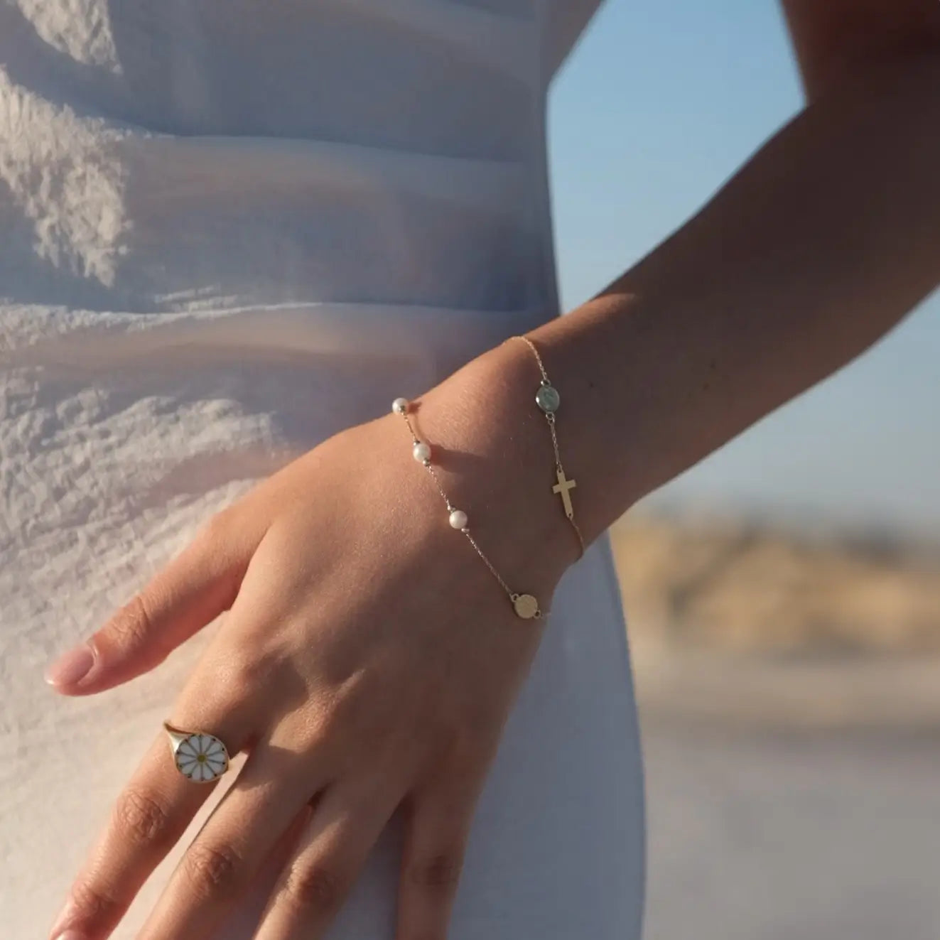 A woman's left hand is adorned with a gold rosary bracelet featuring small pearls and a cross charm. She also wears a ring with a white and gold floral design on her index finger. She is dressed in a light-colored, textured fabric. The background shows a bright outdoor setting with blurred elements.