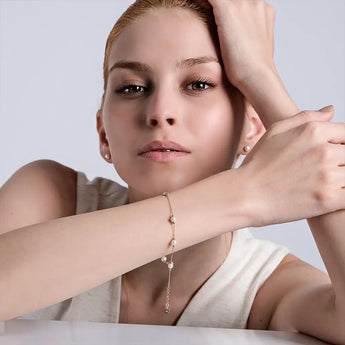 A young woman with light-colored hair leans forward with her arms resting in front of her. She wears a sleeveless light-colored top and a gold rosary bracelet with small pearls on her left wrist. Her expression is calm and contemplative, and she has pearl stud earrings. The background is plain and light-colored.