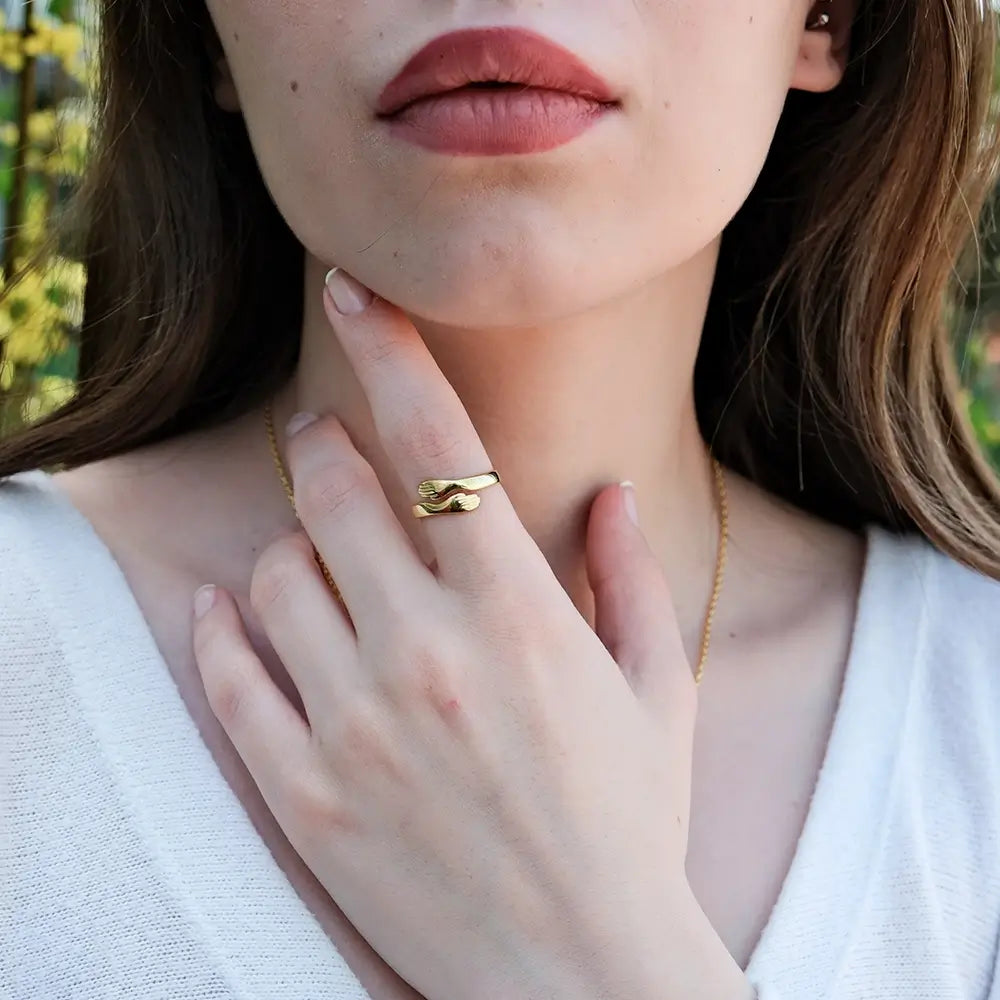 A young woman with light brown hair touches her chin with her left hand, wearing a gold snake-shaped ring on her index finger. She has light pink lips and wears a white top. A delicate gold chain necklace is also visible around her neck. The background is an outdoor setting with blurred green foliage.