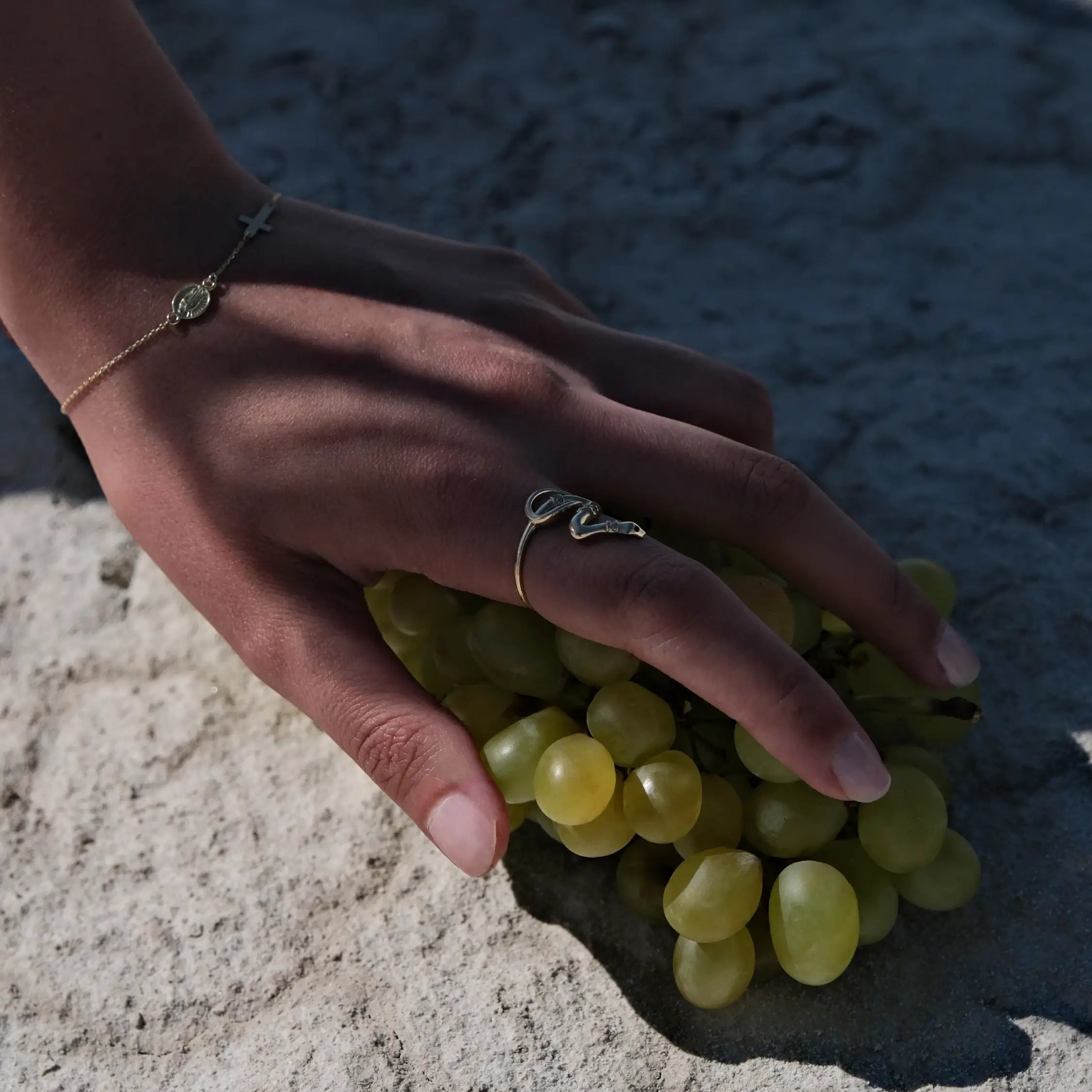 A woman's hand rests on a cluster of green grapes. She wears a gold rosary bracelet on her left wrist and a snake-shaped ring on her left index finger. The background is a textured, light-colored surface.