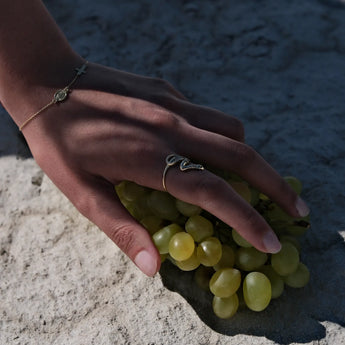 A woman's hand rests on a cluster of green grapes. She wears a gold rosary bracelet on her left wrist and a snake-shaped ring on her left index finger. The background is a textured, light-colored surface.