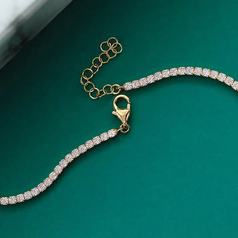 A gold chain bracelet adorned with small, sparkling white gemstones is laid out on a green surface. The bracelet features a lobster clasp and an adjustable extender chain. The background includes a portion of a white and gray marble surface in the top left corner.