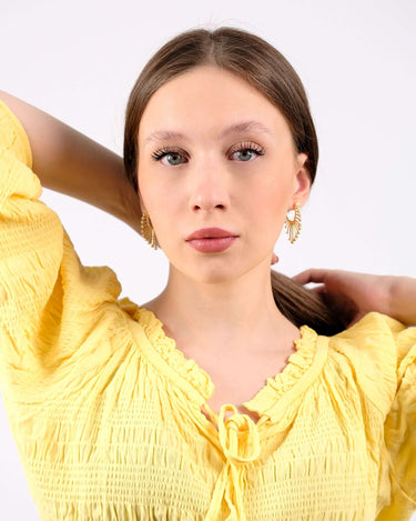 Young woman in a yellow ruffled blouse wearing ornate 14K gold vermeil sunburst earrings, looking directly at the camera with a neutral expression.