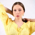 Young woman in a yellow ruffled blouse wearing ornate 14K gold vermeil sunburst earrings, looking directly at the camera with a neutral expression.