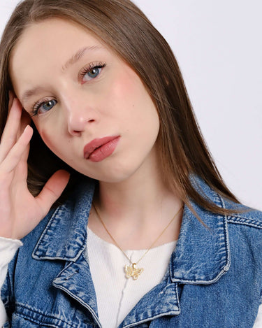 Portrait of a young woman with fair skin and long brown hair, wearing a delicate gold butterfly necklace and a denim jacket, resting her face on her hand.