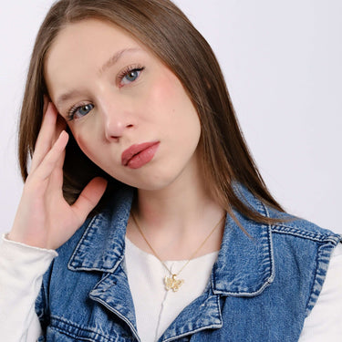 Portrait of a young woman with fair skin and long brown hair, wearing a delicate gold butterfly necklace and a denim jacket, resting her face on her hand.