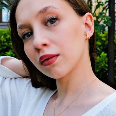 Portrait of a young woman in a white top, wearing 14K gold vermeil X earrings, with a gentle expression and natural setting background.