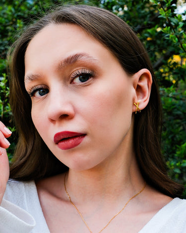 Close-up of a young woman with her hand near her face, showcasing 14K gold vermeil X earrings, set against a garden background.