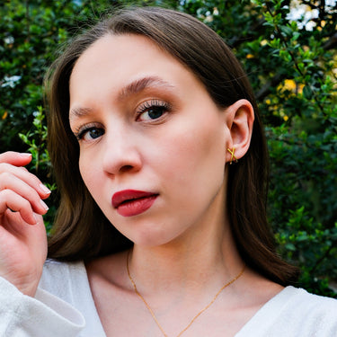 Close-up of a young woman with her hand near her face, showcasing 14K gold vermeil X earrings, set against a garden background.