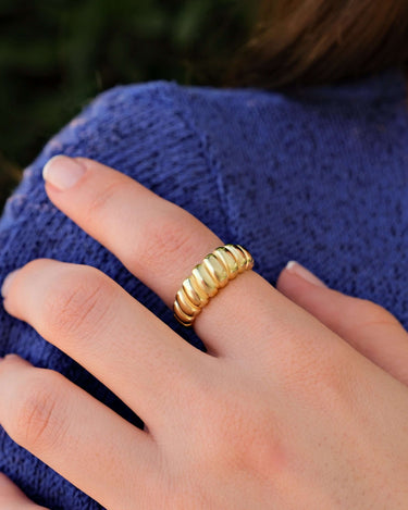 A close-up of a woman&#39;s hand with neatly manicured nails, wearing a 14k gold vermeil croissant ring on her middle finger, resting on a knitted blue sweater.