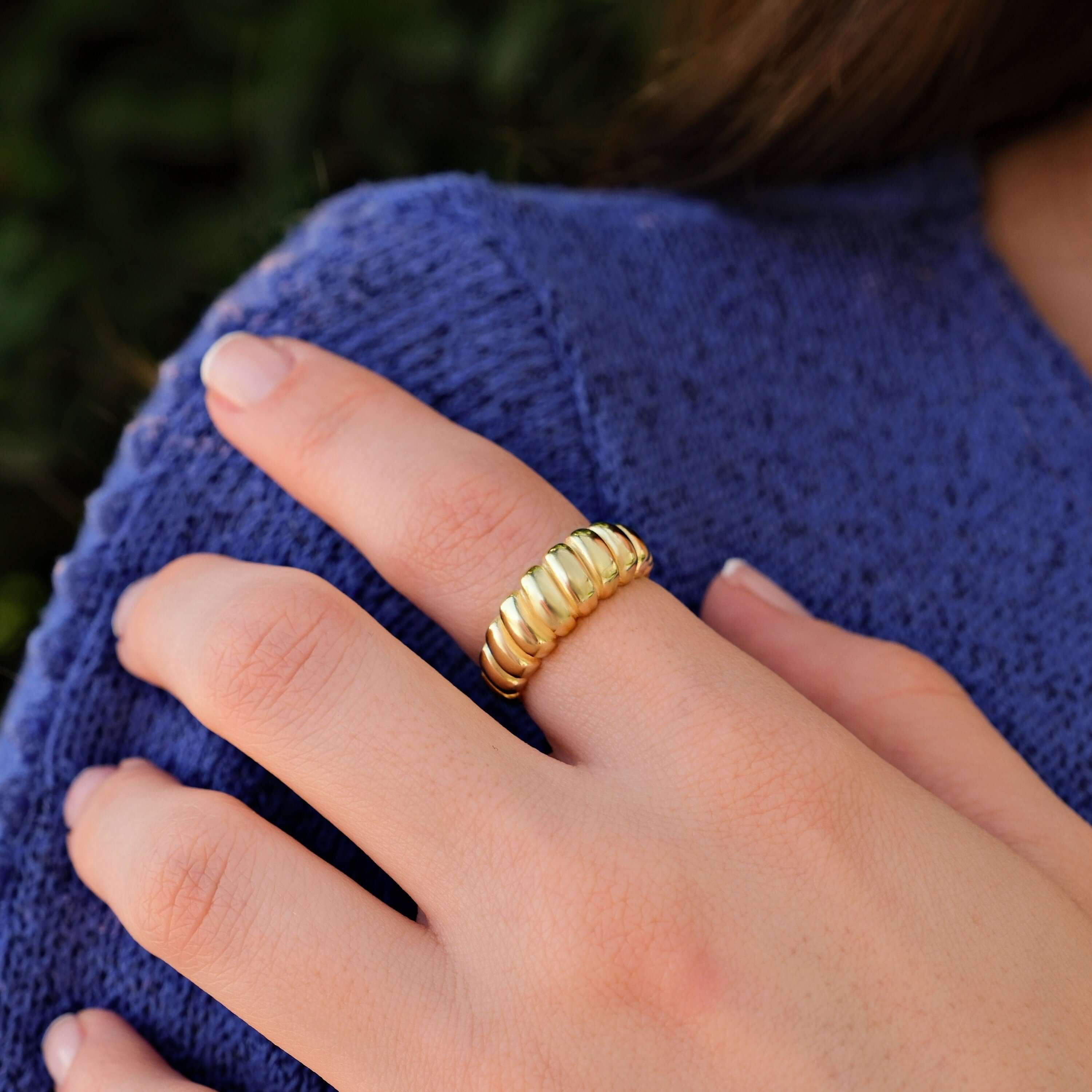 A close-up of a woman's hand with neatly manicured nails, wearing a 14k gold vermeil croissant ring on her middle finger, resting on a knitted blue sweater.