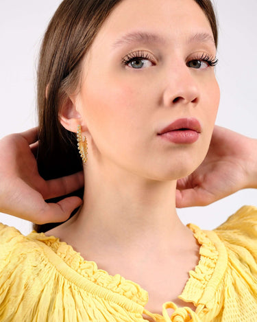 Young woman in a yellow ruffled blouse wearing ornate 14K gold vermeil sunburst earrings, looking directly at the camera with a neutral expression.