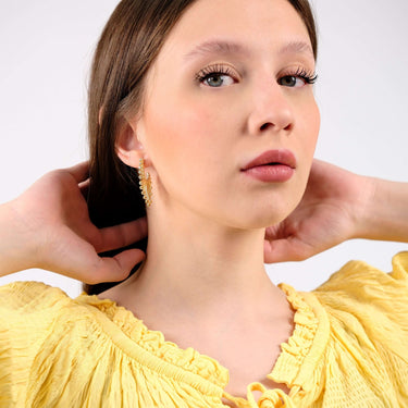 Young woman in a yellow ruffled blouse wearing ornate 14K gold vermeil sunburst earrings, looking directly at the camera with a neutral expression.