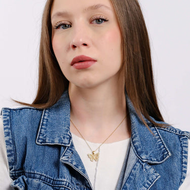 A young woman in a denim jacket and white top pointing subtly at her gold butterfly necklace.