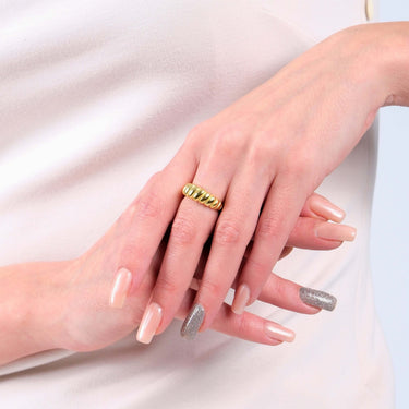 A woman&#39;s hands with manicured nails, one nail painted silver, wearing a 14k gold vermeil croissant ring on her right ring finger, against a light beige dress.