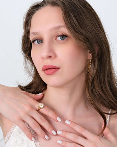 A woman with long brown hair and blue eyes posing with her hands near her face, highlighting the 14K solid gold daisy ring on her finger.