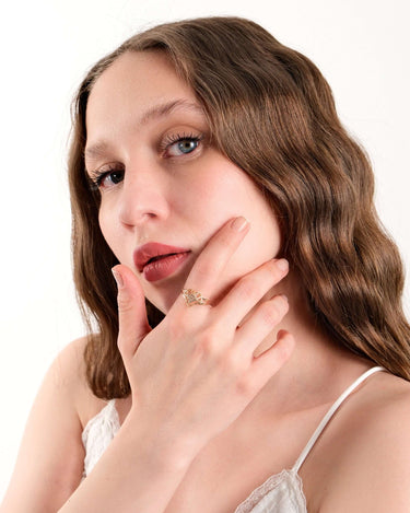 Close-up of a woman with wavy brown hair and blue eyes, touching her chin and displaying a gold heart ring on her finger.