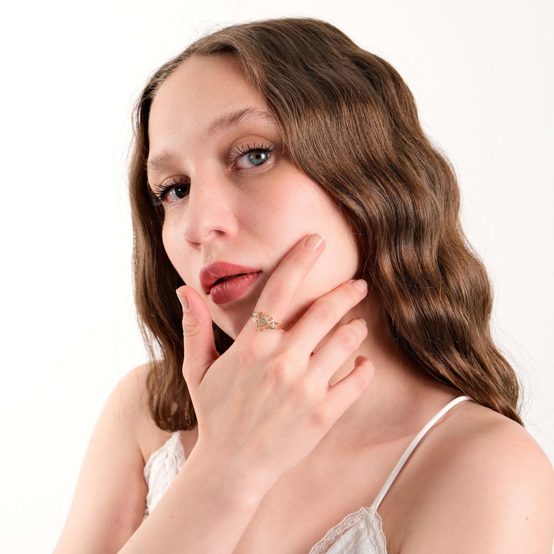 Close-up of a woman with wavy brown hair and blue eyes, touching her chin and displaying a gold heart ring on her finger.