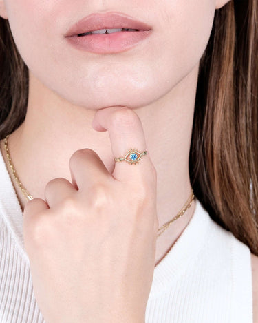 : A close-up of a woman&#39;s face, focusing on her lips and chin. She is lightly touching her lips with her hand, which is adorned with a gold evil eye ring featuring a blue gemstone.