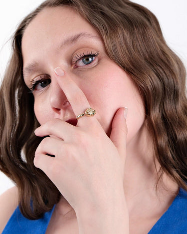 Woman in a blue top touching her face, wearing a gold Ouroboros thumb ring with green gemstones.