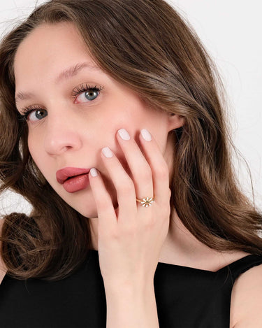 Portrait of a woman with light brown hair, touching her face with a hand adorned with a gold flower ring, wearing a black top.