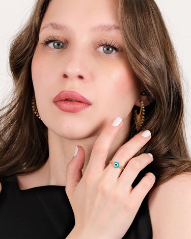 A woman with light brown hair and blue eyes wearing a black top, posing with her hand near her face to display a gold chain ring featuring a hexagonal evil eye design